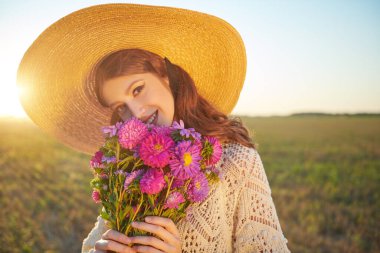 Yaz güzeli kız. Zarif, geniş kenarlı şapkalı romantik bir kızın portresi elinde bir buket çiçekle yaz manzarasının arka planında. Makyaj ve kozmetik.