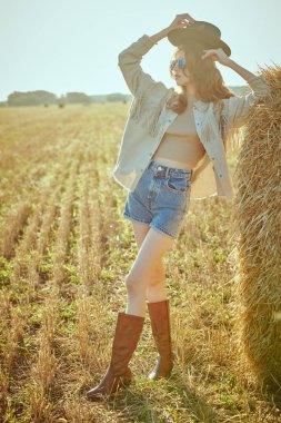 Modern hippi ve kot tarzı. Güzel manken kız, batan güneşin altında samanlıkta samanlığın yanında kot elbise, şapka ve deri botlarla poz veriyor. Yaz ve sonbahar modası.