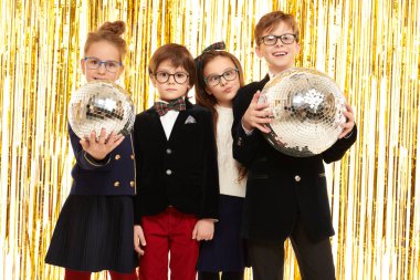 Zarif gece kıyafetleri içindeki mutlu çocuklar ellerinde disko toplarıyla parlak altın folyo perdelerin arkasında neşeyle poz veriyorlar. Çocuk partisi. Çocuk modası ve tatiller. 