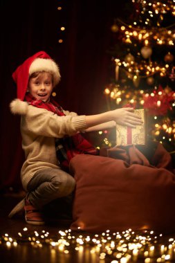 Noel Baba şapkalı neşeli çocuk evde çantasından hediye alıyor. Noel arkaplanı ve etrafta parlayan ışıklar. Noel Arifesi.