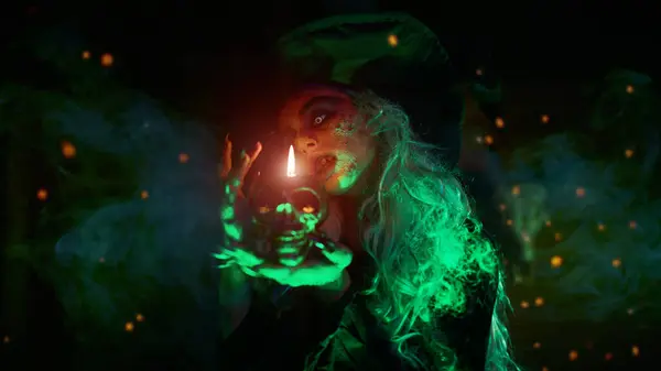 stock image A scary wicked witch conjures in her lair and holds a skull candle in her hands. Magical green light and sparks of fire around her. Halloween. 