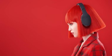 Kırmızı dudaklı, kızıl saçlı, kırmızı kareli ceket ve kulaklık takan şık bir kızın yan görüntüsü. Fotokopi alanı olan kırmızı stüdyo. DJ, parti kızı. Güzellik, Moda. 