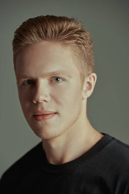 Kameraya gülümseyen siyah tişörtlü yakışıklı bir sarışının portresi. Gri arka plan. Gençler. 