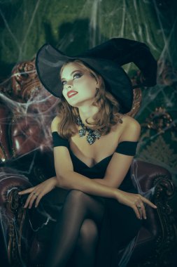 Cadılar Bayramı Cadısı. Şapkalı ve omuzları açık güzel genç bir cadı eski şatoda örümcek ağıyla kaplı eski bir koltukta rüya gibi oturuyor. Cadılar Bayramı için peri masalları. 