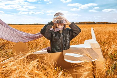 Deri ceketli ve havacı kasklı neşeli bir çocuk dürbünle uzaktaki karton bir uçakta oturuyor. Buğday tarlası arka planı güneş ışığıyla aydınlatılıyor. Çocuk oyunları ve hayalleri. 