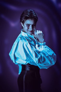 Zarif 19. yüzyıl kıyafetleri içinde çekici bir genç vampir kameraya davetkar bir şekilde bakar ve sinsice gülümser ve dişlerini açığa çıkarır. Siyah ve mor arkaplanda stüdyo portresi. Cadılar Bayramı.