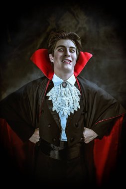 Vampir dünyası. Siyah paltolu ve pelerinli komik ve korkutucu genç bir vampirin portresi büyük gözlerle kameraya bakıyor ve şeytanca gülüyor. Cadılar Bayramı. 