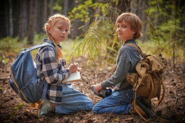 Aktif çocuklar. Sırt çantalı iki meraklı çocuk ormanda doğa gezileri sırasında not defterlerine not alıyorlar. Çocuklar için macera ve doğa keşfi. 