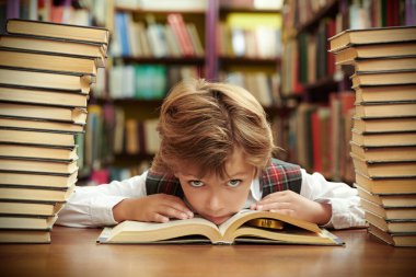 Üniformalı yorgun bir öğrenci, bir okul kütüphanesinde çenesi bir kitaba dayanmış kitap yığınlarının arasında oturur. Modern eğitim ve çocuklar.
