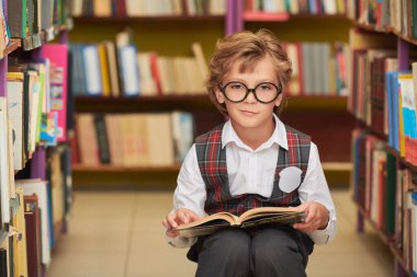 Temiz okul üniformalı ve gözlüklü zeki bir çocuk elinde açık bir kitapla kütüphanede oturuyor. Akıllı okul çocuğu. Eğitim ve çocuklar.