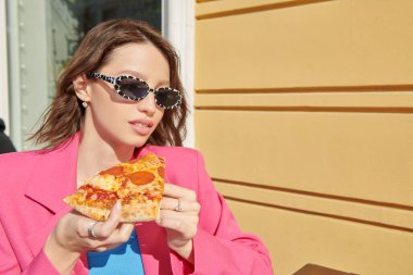 Şık güneş gözlüklü ve parlak yaz elbiseli güzel bir kız açık hava kafede bir dilim pizza yiyor. İnsanlar ve yaşam tarzı.