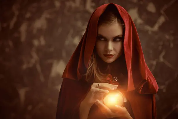 Black magic. A bewitching dark witch woman in a red cape with a hood conjures with a crystal ball in her hands and looks away unkindly. Magic tales. Halloween. Copy space.