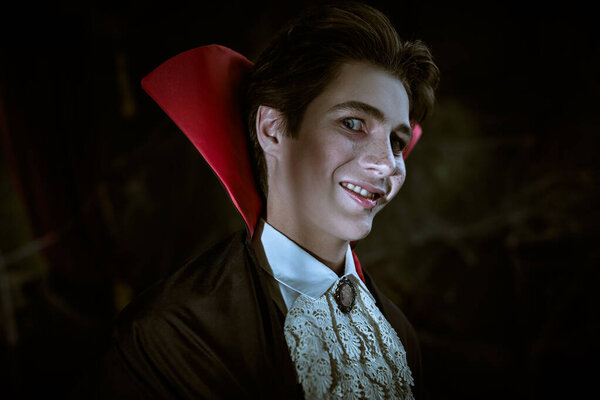 Portrait of a teenage vampire aristocrat in an elegant suit and cloak with a red collar, who looks at the camera and smiles wickedly with drops of blood on his lips. Black background. Halloween.