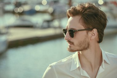 Zarif siyah güneş gözlüklü ve beyaz gömlekli yakışıklı bir adam yat limanında duruyor. Erkek güzelliği ve moda. Denizde yaz tatili. Boşluğu kopyala.