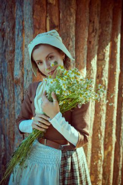 Portrait of a young woman in a Scottish costume of the 18th century standing by wooden fence with a bunch of camomiles. Historical reconstruction of the 18th century. clipart