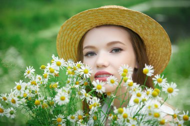 Yazın hasır şapkalı, kahverengi saçlı romantik bir kız bir buket taze papatya çiçeği ve gülümsemeyle bir yaz günü dışarıda duruyor. Yaz güzelliği, makyaj ve çiçekler..