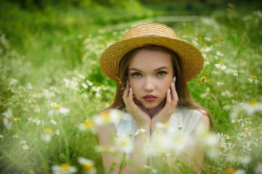 Yaz güzelliği, makyaj ve çiçekler. Yazlık hasır şapkalı güzel kahverengi saçlı bir kız yeşil çimenlerin ve papatyaların arasında oturuyor.. 