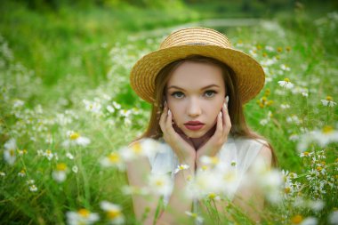 Hasır şapkalı hassas bir kız yeşil çimenler ve papatyalarla çevrili bir şekilde tarlada oturuyor. Yaz güzelliği, makyaj ve çiçekler..