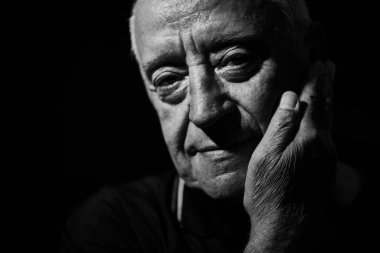 70 'li yaşlarında yaşlı bir adamın elini yanağında tutarken ve sakin bir gülümsemeyle kameraya bakarken yakın çekim. Siyah bir arkaplanda stüdyo resmi.