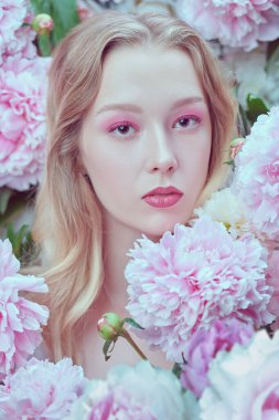 Dalgalı sarı saçlı, zarif pembe makyajlı güzel bir kızın portresi şakayık çiçeklerinin arasında duruyor. Makyaj ve makyaj malzemeleri. Parfüm. Güzel kız.. 