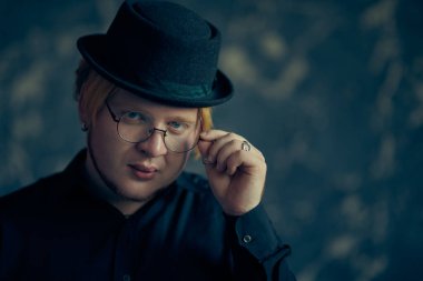 Erkek tarzı. Kırmızı saçlı, tombul yanaklı, siyah tişörtlü, şık siyah şapkalı ve yuvarlak gözlüklü bir genç grunge arka planda poz veriyor. Stüdyo çekimi. Şık bir genç adam. Boşluğu kopyala.