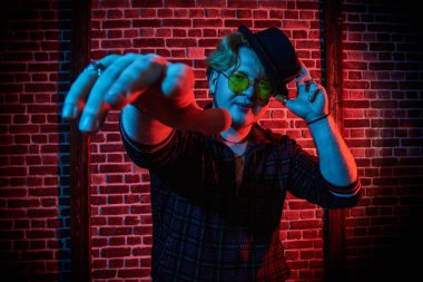 Modern kıyafetlerle, şık siyah şapkalı ve sarı yuvarlak gözlüklü tombul bir rapçi portresi koyu bir grunge arka planda sokak dansı yapıyor. Gece kulübündeki rapçi çocuk. Erkek gençlik tarzı.