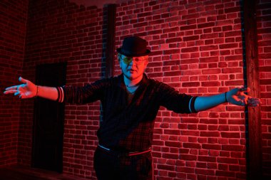 Modern kıyafetlerle, şık siyah şapkalı, sarı yuvarlak gözlüklü, kırmızı ışıkla aydınlatılmış çatı katında dans eden rapçi bir adamın portresi. Gece kulübündeki rapçi çocuk. Erkek gençlik tarzı.