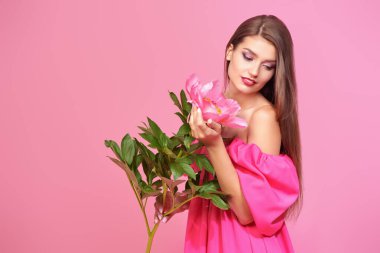 Uzun kahverengi saçlı, omzundan pembe elbisesi olan sevimli bir kız pembe şakayık çiçeğiyle poz veriyor. Nazik pembe arka plan. Güzellik ve çiçekler, makyaj. Boşluğu kopyala.