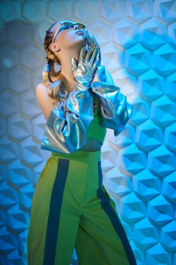 Özel tasarım yaz tarzı. Moda tasarımcısı kıyafetleri, eldivenleri ve güneş gözlükleriyle mavi ışık altında güzel bir kadın model poz veriyor. Yüksek moda. 