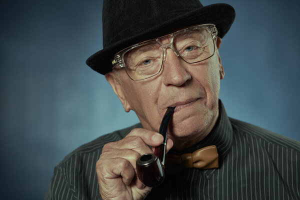 Старый хорошо одетый джентльмен, курящий трубку. Студийный портрет на темно-синем фоне.