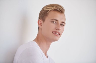 Yakışıklı genç sarışın bir adam beyaz bir duvara yaslanmış ve gülümsüyor. Mutlu insanlar, olumlu duygular. Erkek güzelliği ve sağlığı. 