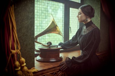 Adorable aristocratic girl in a black elegant dress with dark gathered hair stands by the window in a vintage room listening to the gramophone. The heroine of the novel, 19th century style. clipart