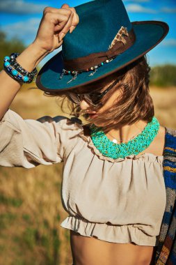 Kadınsı bir bluz, şapka ve güneş gözlüğü takmış çekici bir boho kızı güneşli bir günde bir tarlanın ve mavi gökyüzünün arkasında poz verir. Yaz tatili, seyahat. Bohem, modern hippi tarzı.. 