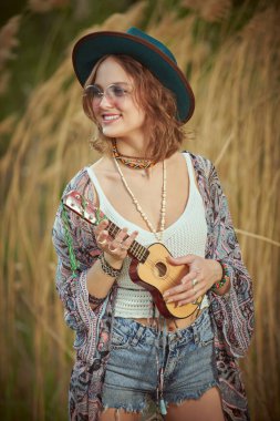 Boho tarzı kıyafetler ve aksesuarlarla dolu neşeli bir kız sazlıkların yanındaki bir tarlada durup neşeyle gülümsüyor ve ukulele çalıyor. Boho ve hippi tarzı. Yaz, tatil.