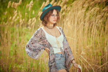 Boho tarzı elbiseleri ve aksesuarları olan şık, güzel bir kız sazlıkların yanındaki bir tarlada duruyor. Boho ve hippi tarzı. Tatil, seyahat.