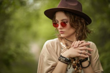 Boho tarzı kıyafetler ve güneş gözlüğü ve şapka takan güzel bir kız yaz parkında yeşil ağaçların arkasında duruyor. Moda fotoğrafı. Boho ve hippi tarzı.