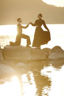 Genç yakışıklı adam gün batımında deniz kıyısında duran güzel genç bir kadının önünde diz çöktü. Tarihi aşk romanı. Sevgi dolu bir ilişki. 19. yüzyıl tarzı.