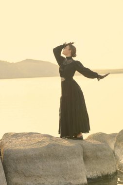 Siyah zarif elbiseli zarif bir bayan, ne yazık ki deniz kenarında duygularından muzdarip duruyor. 19. yüzyıl romanı.