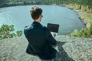 Siyah takım elbiseli başarılı bir iş adamı güzel manzarayla çevrili bir dizüstü bilgisayarla çalışır. İş hayatında başarı, uzaktan çalışma. Tatil ve seyahat.