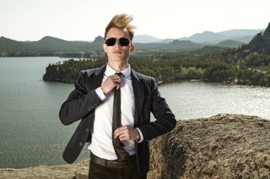İş stili. Katı takım elbiseli, siyah güneş gözlüklü yakışıklı bir adam bir dağ manzarası ve gölün arka planında duruyor. İş, başarı. Tatil.