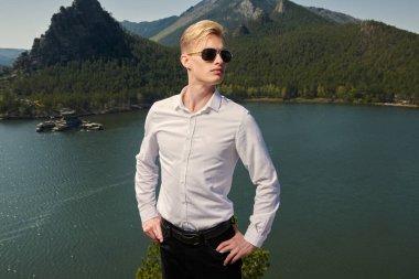 Beyaz gömlekli ve siyah güneş gözlüklü kasıtlı genç bir adam bir göle ve dağ manzarasına karşı duruyor. İş konsepti. Tatil ve seyahat.