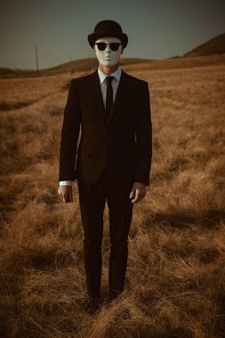 Surreal masked gentleman. Full-length portrait of a man in a black suit, bowler hat and white mask, standing in a field. clipart