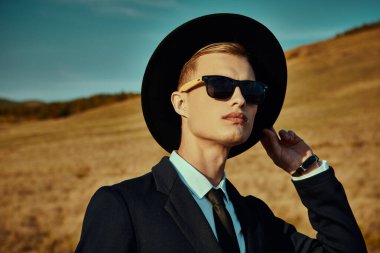 İş stili. Siyah takım elbiseli, beyaz gömlekli, siyah şapkalı ve güneş gözlüklü yakışıklı sarışın bir adam bir vadide duruyor. Moda fotoğrafı.. 