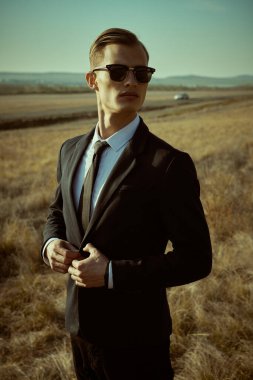 Mükemmel bir adamın portresi. Siyah takım elbiseli, beyaz gömlekli ve siyah güneş gözlüklü, tepelik bir vadide duran yakışıklı sarışın bir adamın moda fotoğrafı. İş biçimi. 