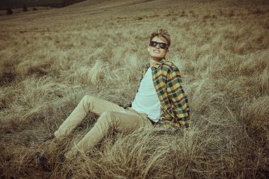 Yakışıklı genç adam kuru çimenlerin üzerinde rahatça oturup bozkırda özgürlüğün tadını çıkarıyor. Erkek modası. 