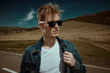 Kot elbiseli, siyah güneş gözlüklü yakışıklı otostopçu resimli bir manzaranın üzerinde duruyor. Denim modası. Yol maceraları, seyahat.