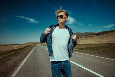 Kot pantolon ve siyah güneş gözlüklü çekici bir otostopçu mavi bir gökyüzü ve vadinin arkasındaki yolda duruyor. Denim modası. Yol maceraları, seyahat.
