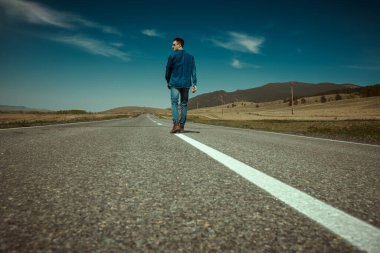 Kot pantolon ve siyah güneş gözlüğü takmış otostopçu bir gencin portresi mavi bir gökyüzü ve vadide otobanda yürüyor. Denim modası. Yol maceraları, seyahat.