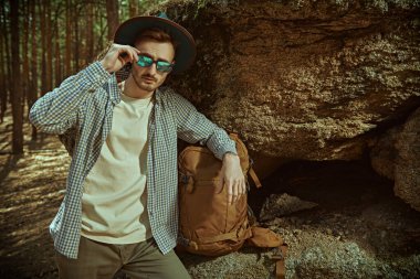 Aktif dinlenme, yürüyüş, macera. Sırt çantasıyla ormanda büyük bir kayanın yanında duran genç bir adam. Turizm, doğa keşfi. Yaz tatili açık havada.