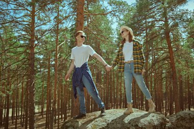 Neşeli bir çift hippi güneşli bir çam ormanında ellerini tutarak yürür. Yaz tatili dışarıda. Turizm. Denim modası. 